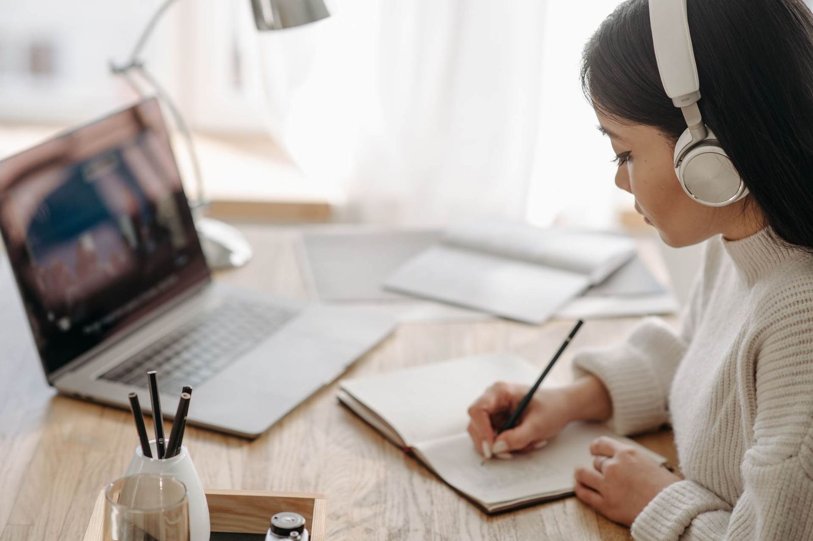 A woman wearing headphones writes in a notebook beside an open laptop at a desk