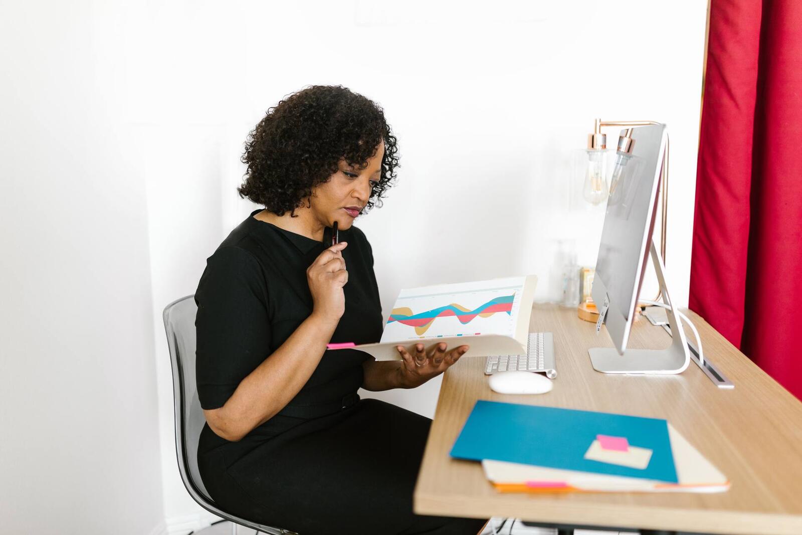 A woman in a black dress reads a folder of colored charts at her desk, pen near her chin