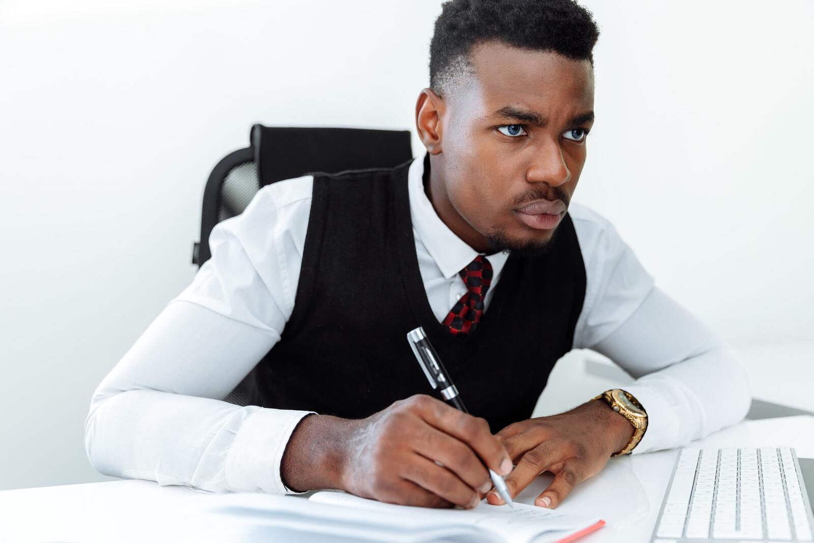 A man in a black sweater vest and tie writes in a notebook at a white desk, keyboard visible beside him, looking off to the side in thought