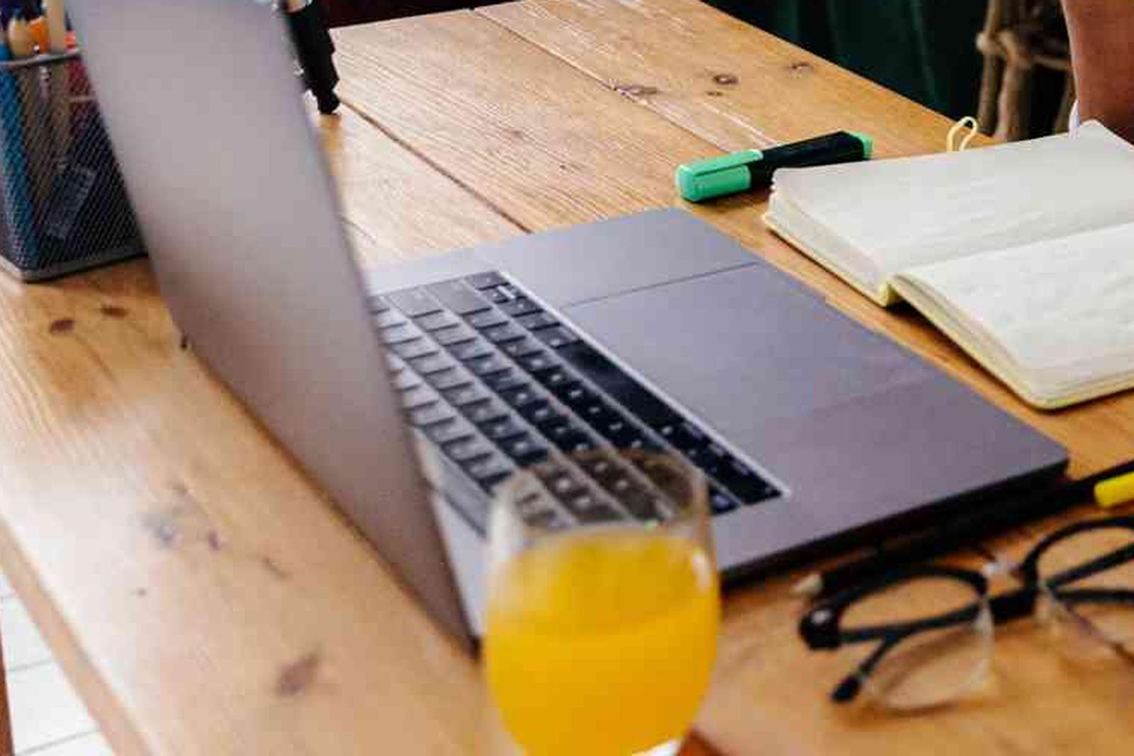 An open laptop and notebook on a wooden desk beside a pair of glasses