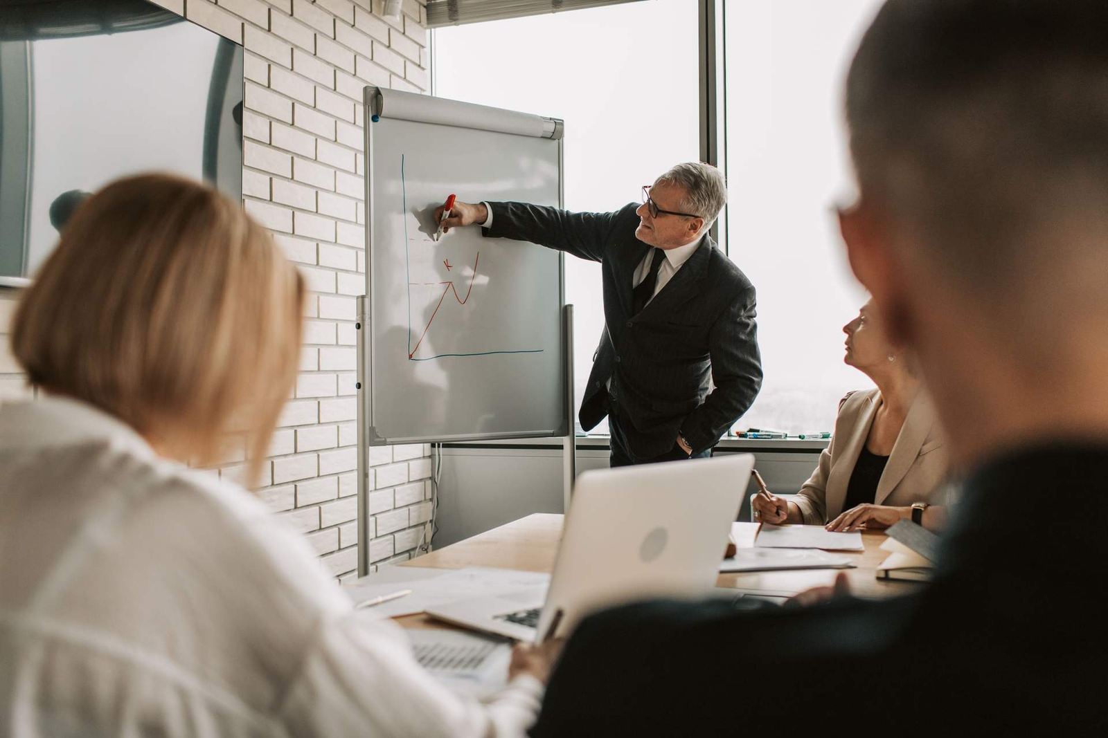 A gray-haired man in a suit points to a hand-drawn chart on a flip chart while colleagues look on in a bright meeting room