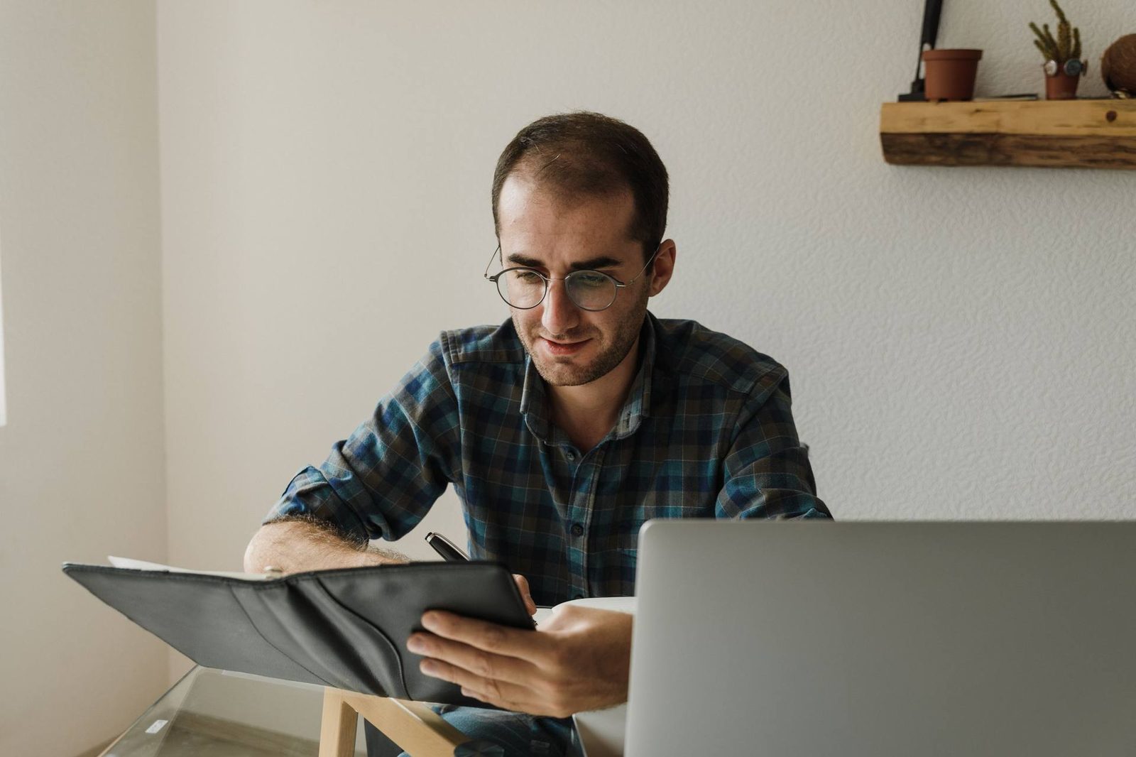 A man at a home desk reviews a document in an open folder while writing, with a laptop open in front of him