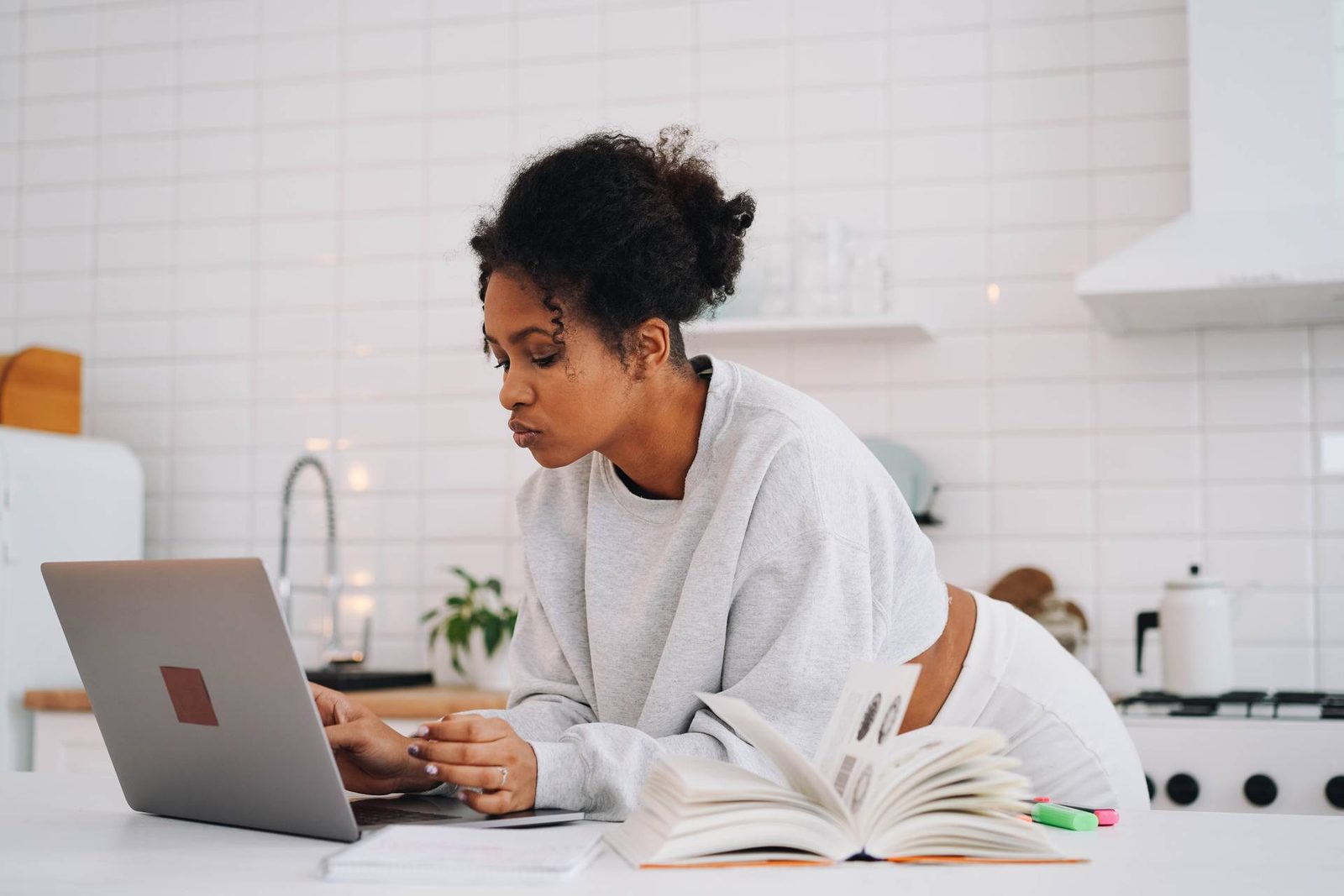 A woman leans over a laptop at a kitchen counter beside an open book