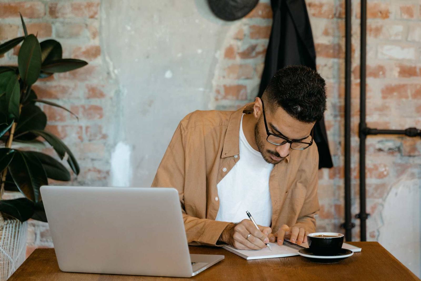 A man wearing glasses writes in a notebook beside an open laptop and a cup of coffee