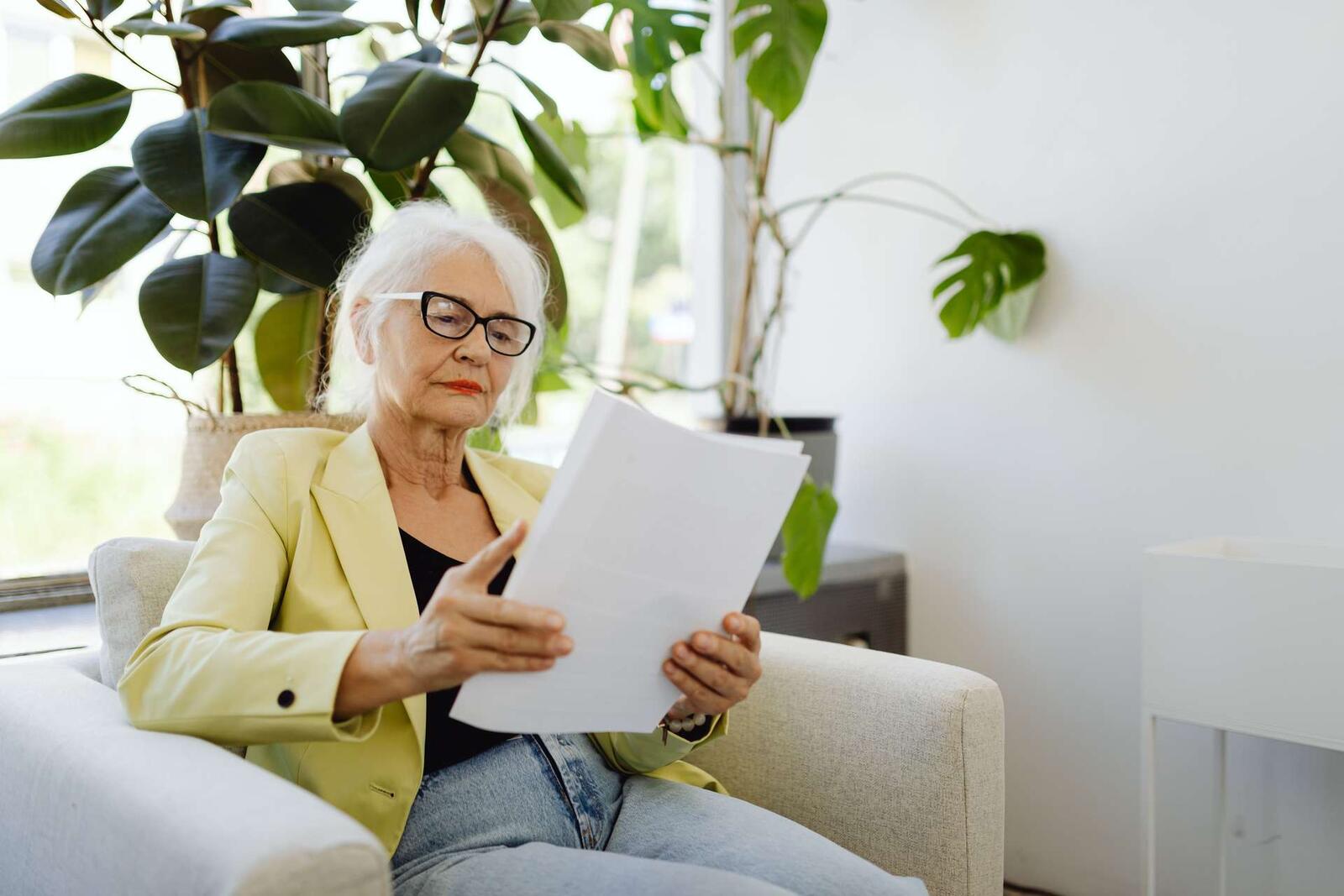 An older woman in a yellow blazer and glasses reads a stack of papers in an armchair surrounded by houseplants