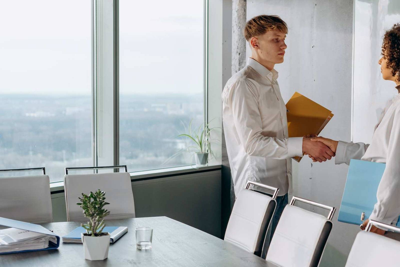 Two colleagues shake hands over an exchanged folder in a conference room with a city view