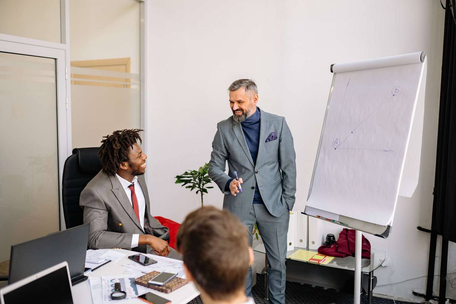 Three colleagues talk and smile around a desk in an office, with a flip chart showing a simple diagram in the background