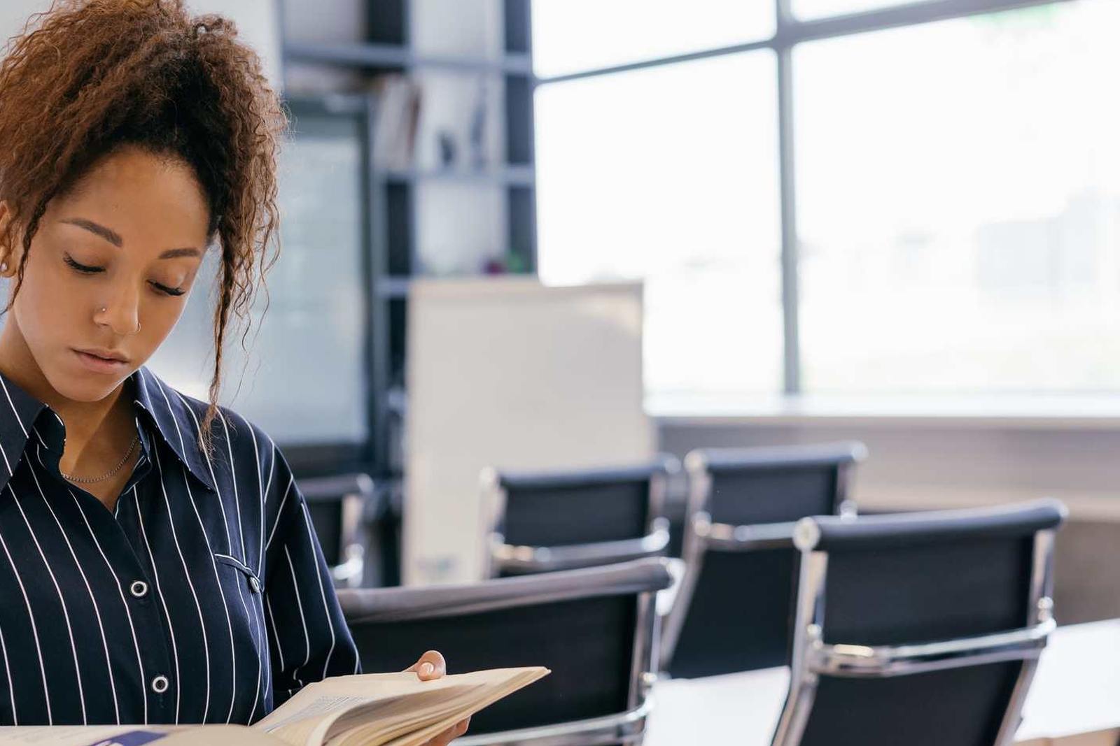 A woman reads closely from an open book while standing in an empty conference room