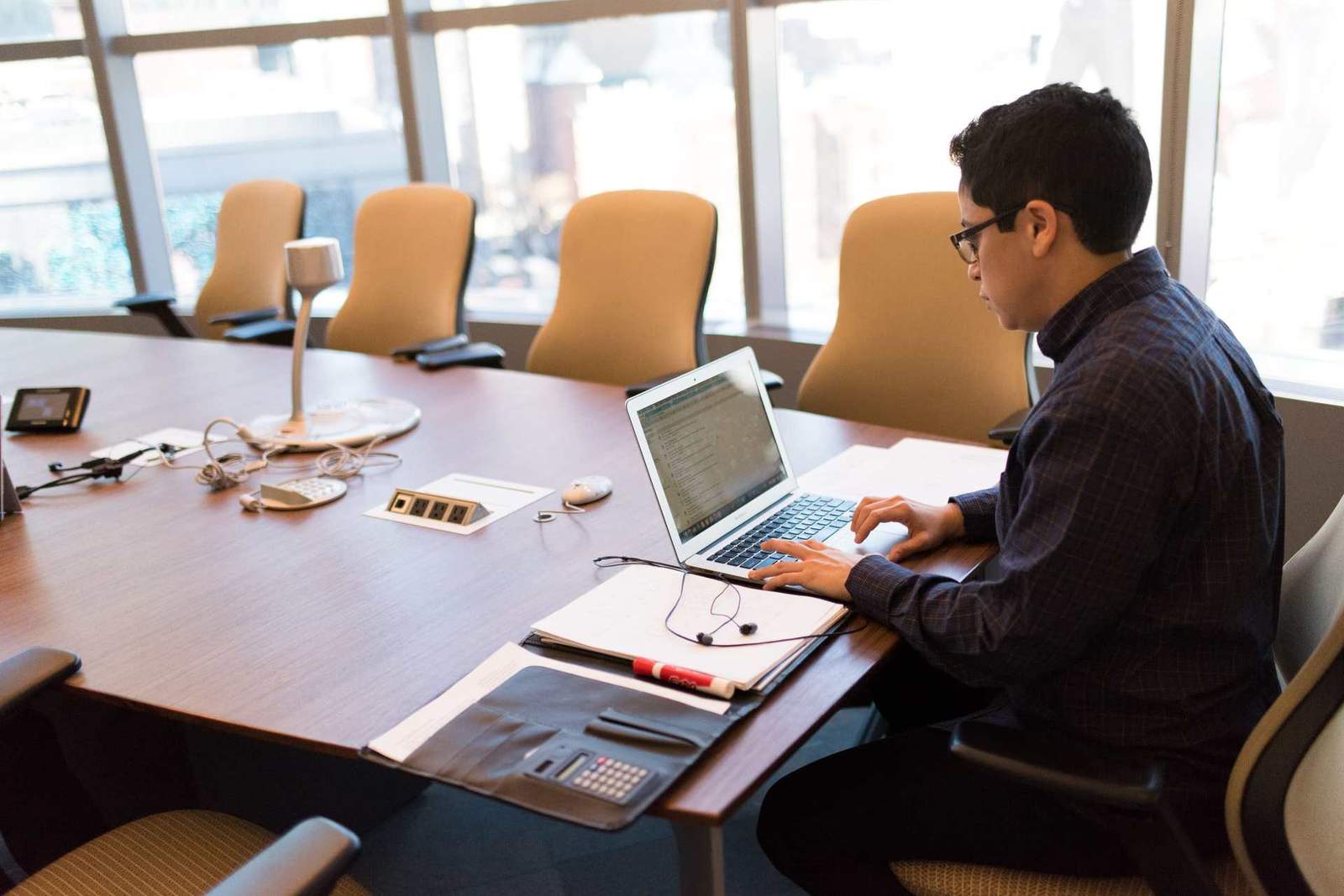 A man works intently on a laptop at an empty conference table, papers and earbuds within reach