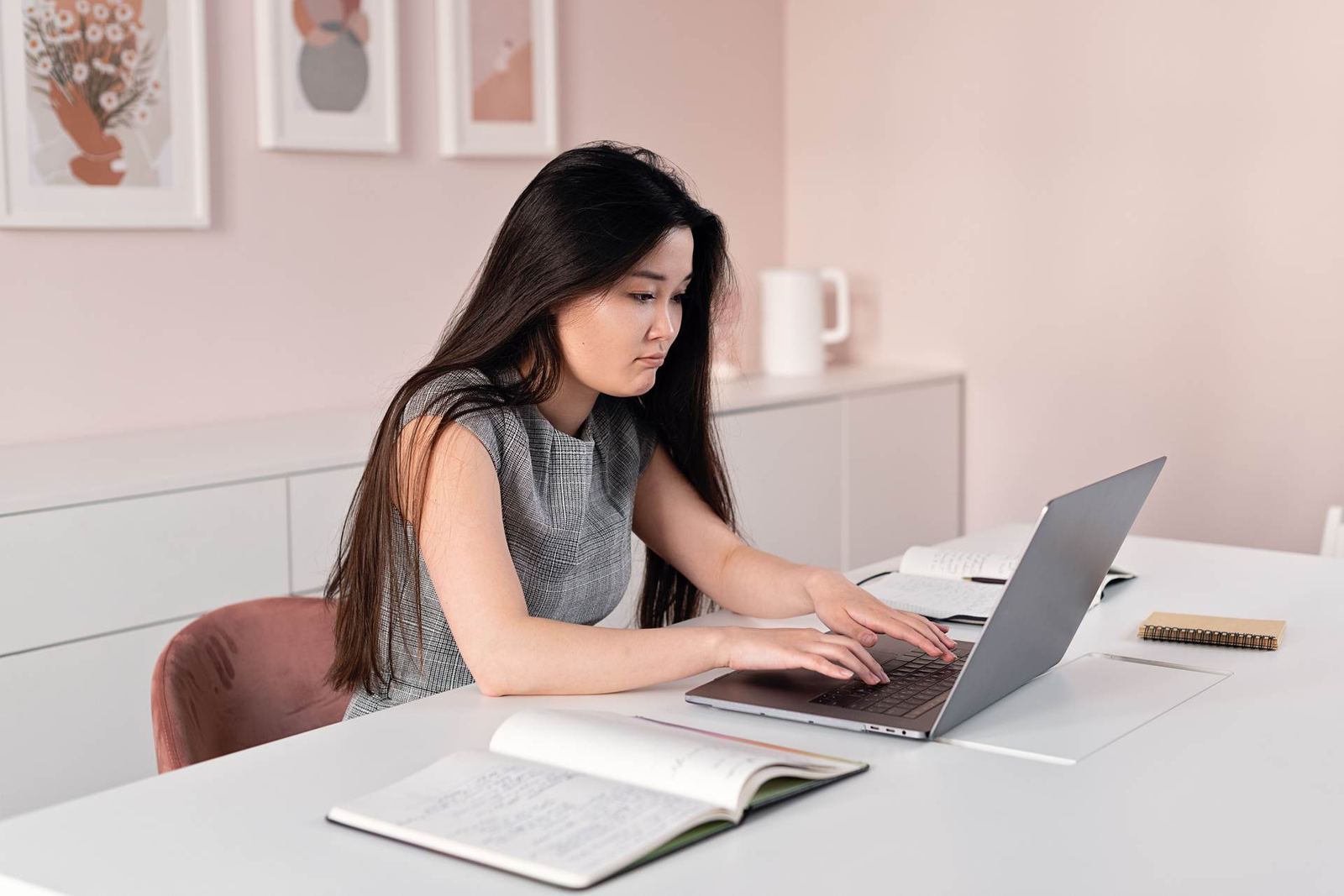 A woman types on a laptop at a desk with open handwritten notebooks in front of her
