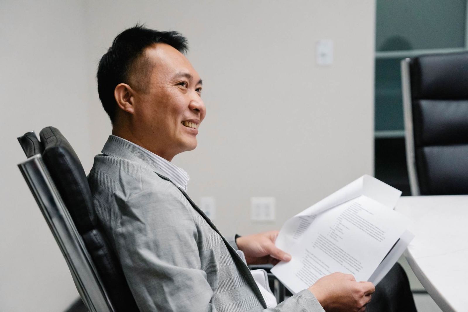 A man in a gray suit sits in an office chair, smiling while reading a printed letter