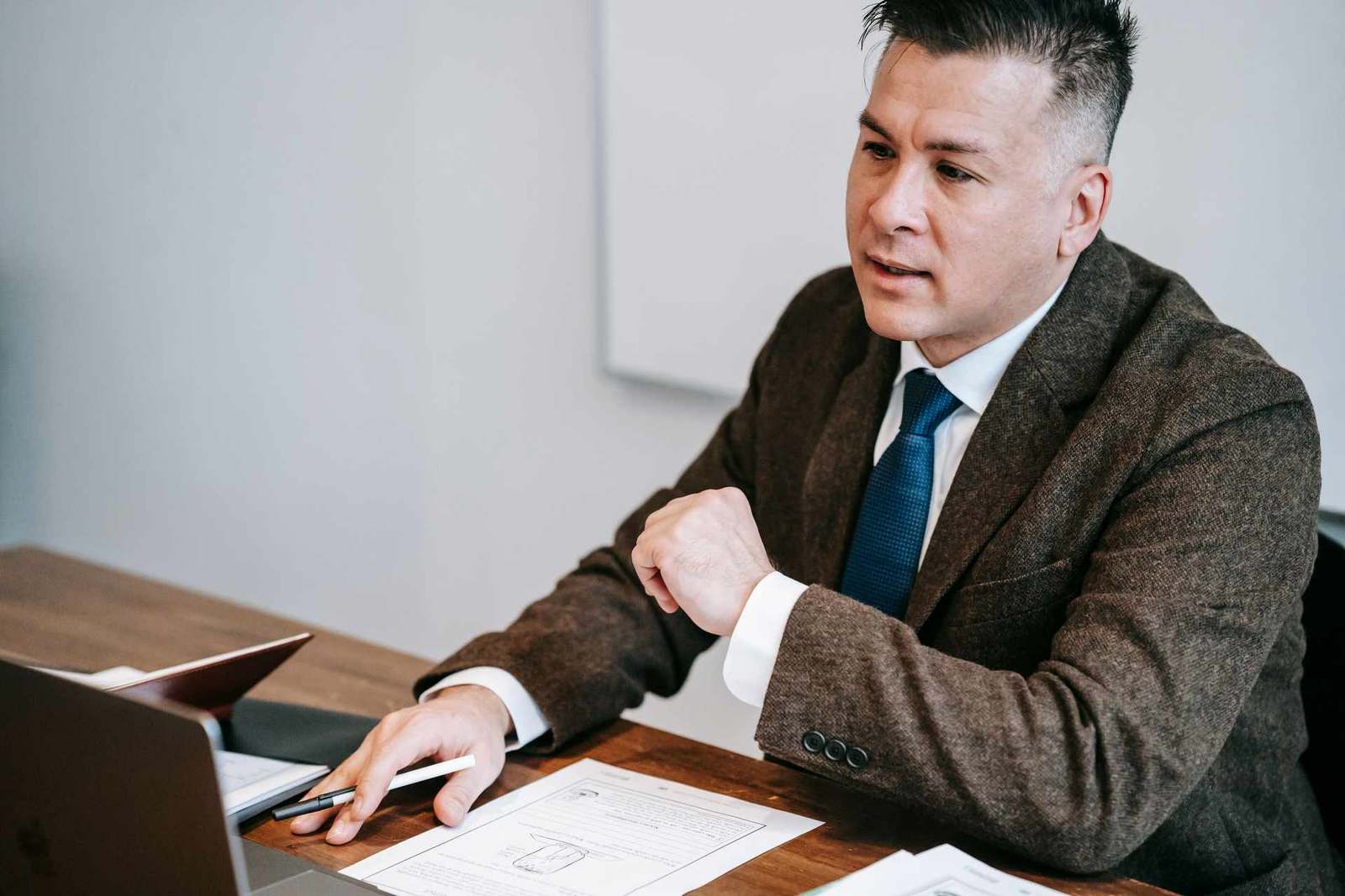 A man in a tweed blazer reviews a printed document at his desk, pen in hand, laptop open beside him