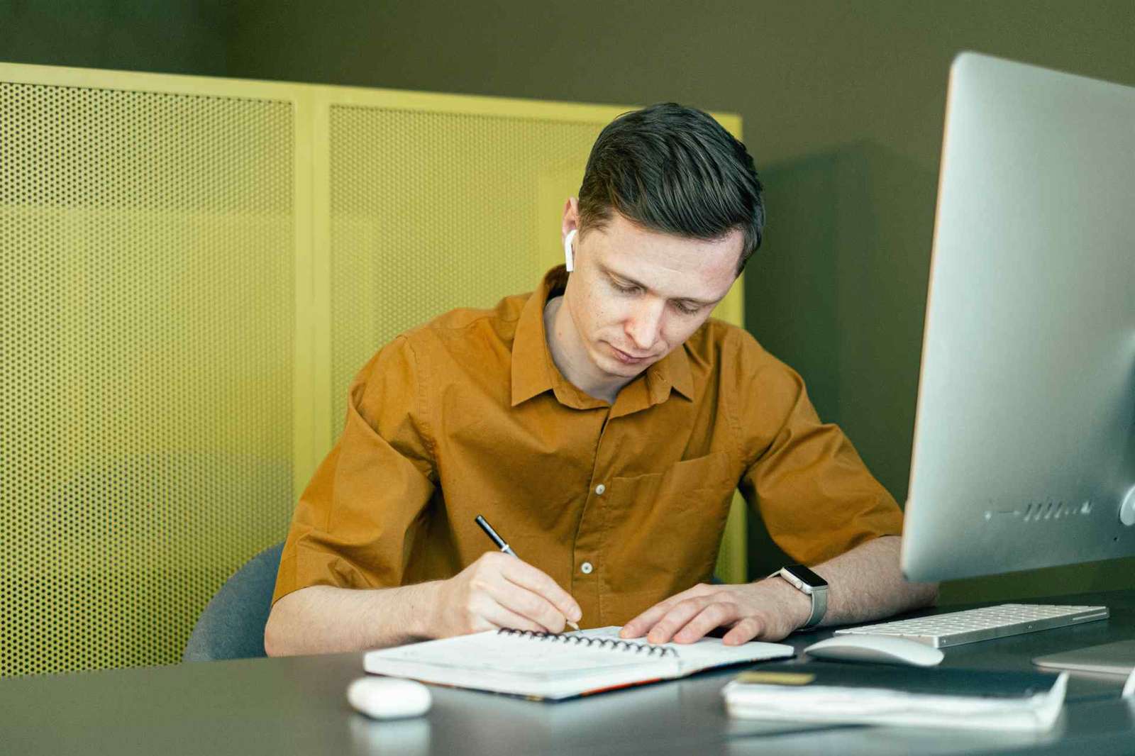 A man writes notes in a spiral notebook at an office desk beside a desktop monitor and keyboard