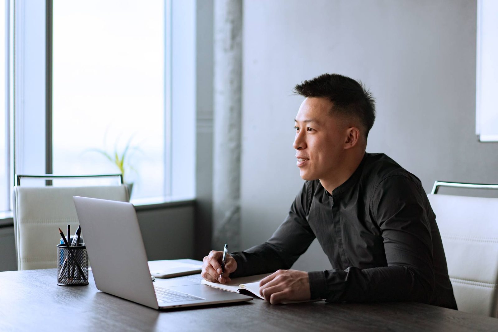 A man in a dark shirt smiles while taking notes at a desk beside an open laptop, looking toward a window