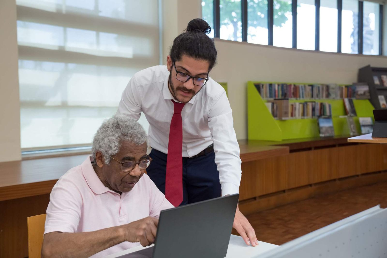 A man in glasses and a red tie leans over to help an older man review a document on a laptop in a library