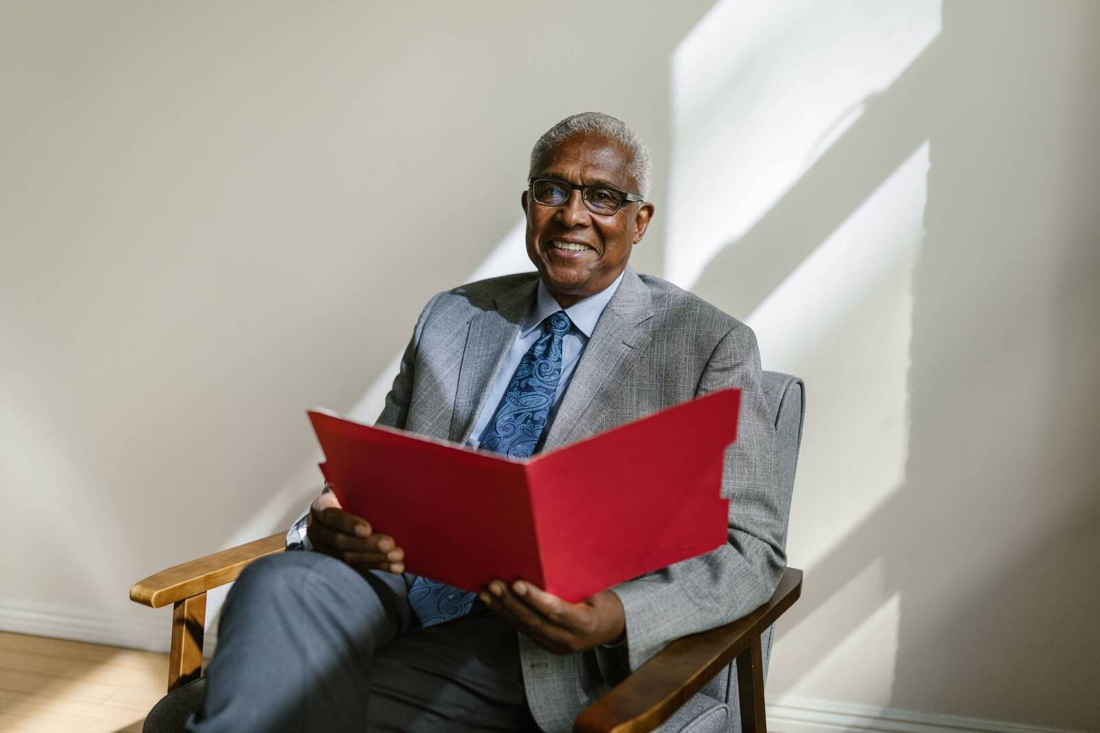 A smiling senior man in a gray suit sits in a sunlit chair holding open a red folder