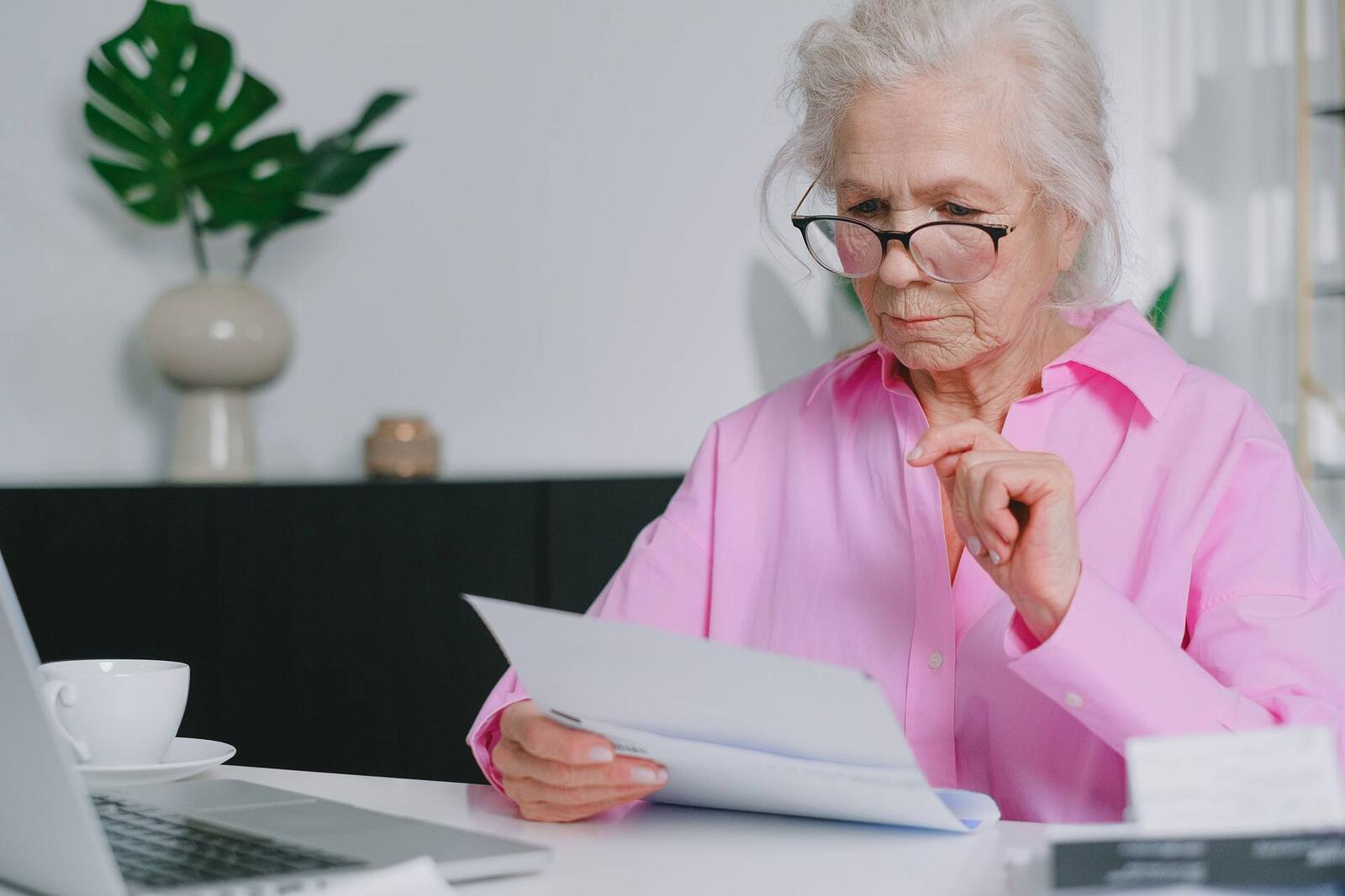 An older woman in a pink shirt and glasses studies a printed letter at her desk, a laptop and coffee cup nearby
