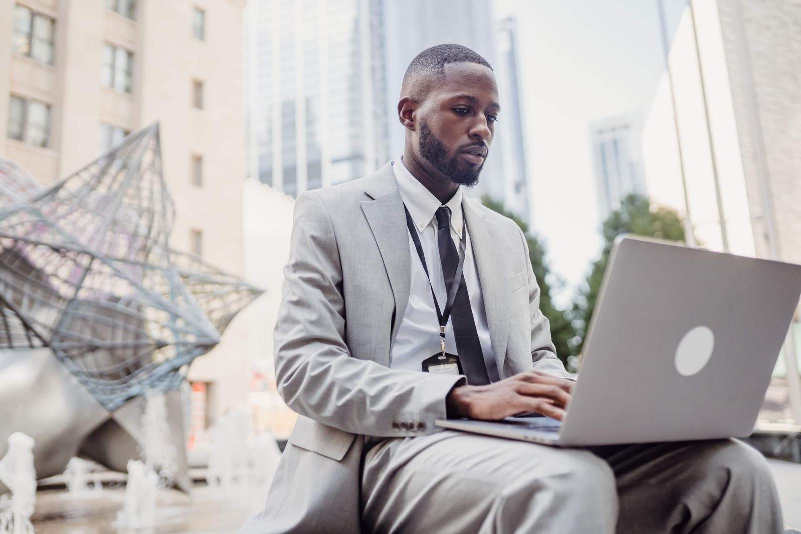 A man in a suit works on a laptop outdoors near modern office towers, an employee badge visible on a lanyard