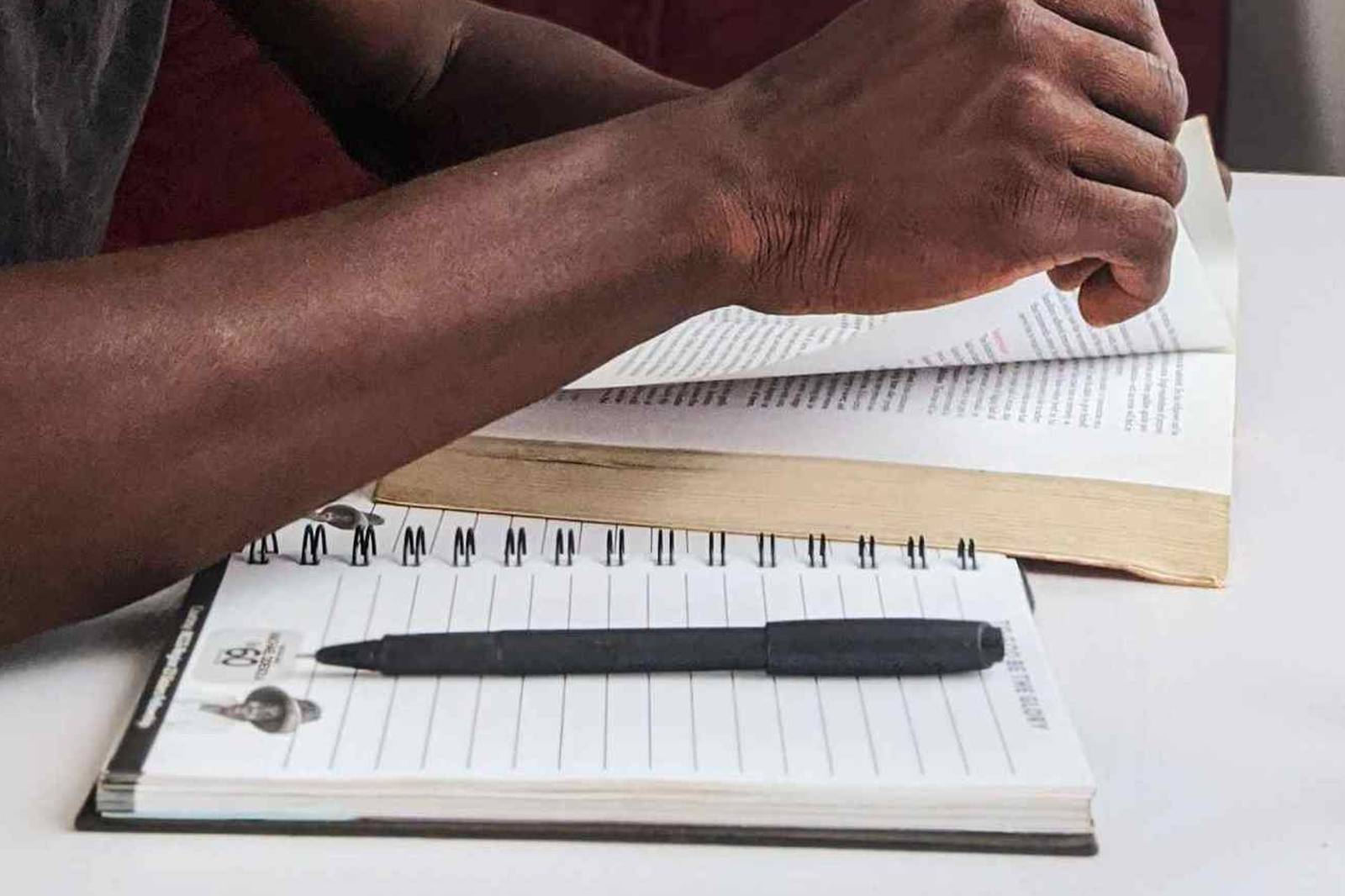 A close-up of a person's hand turning a page in a hardcover book beside a spiral notepad and pen