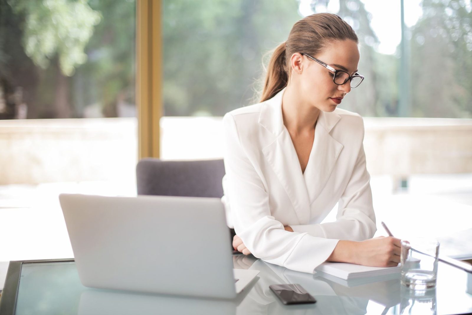 A woman in a white blazer sits at a glass desk writing in a notebook beside an open laptop, with a window in the background