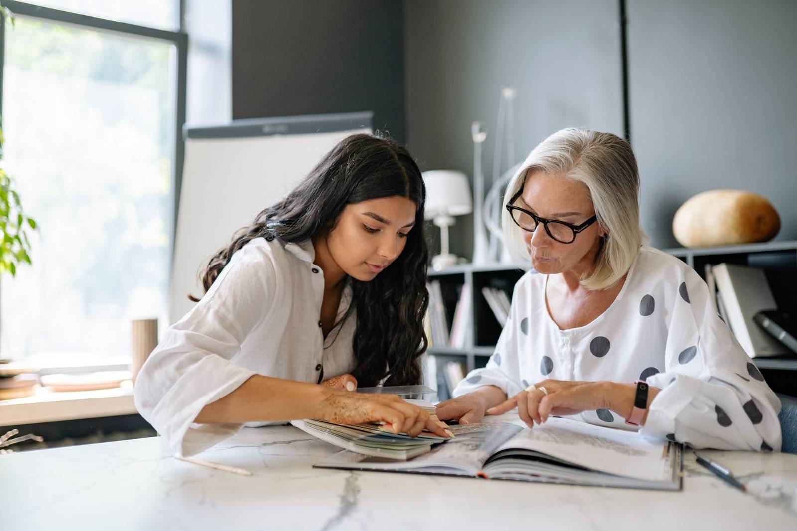 Two women look closely at an open book together at a desk, one pointing at the page while the other listens