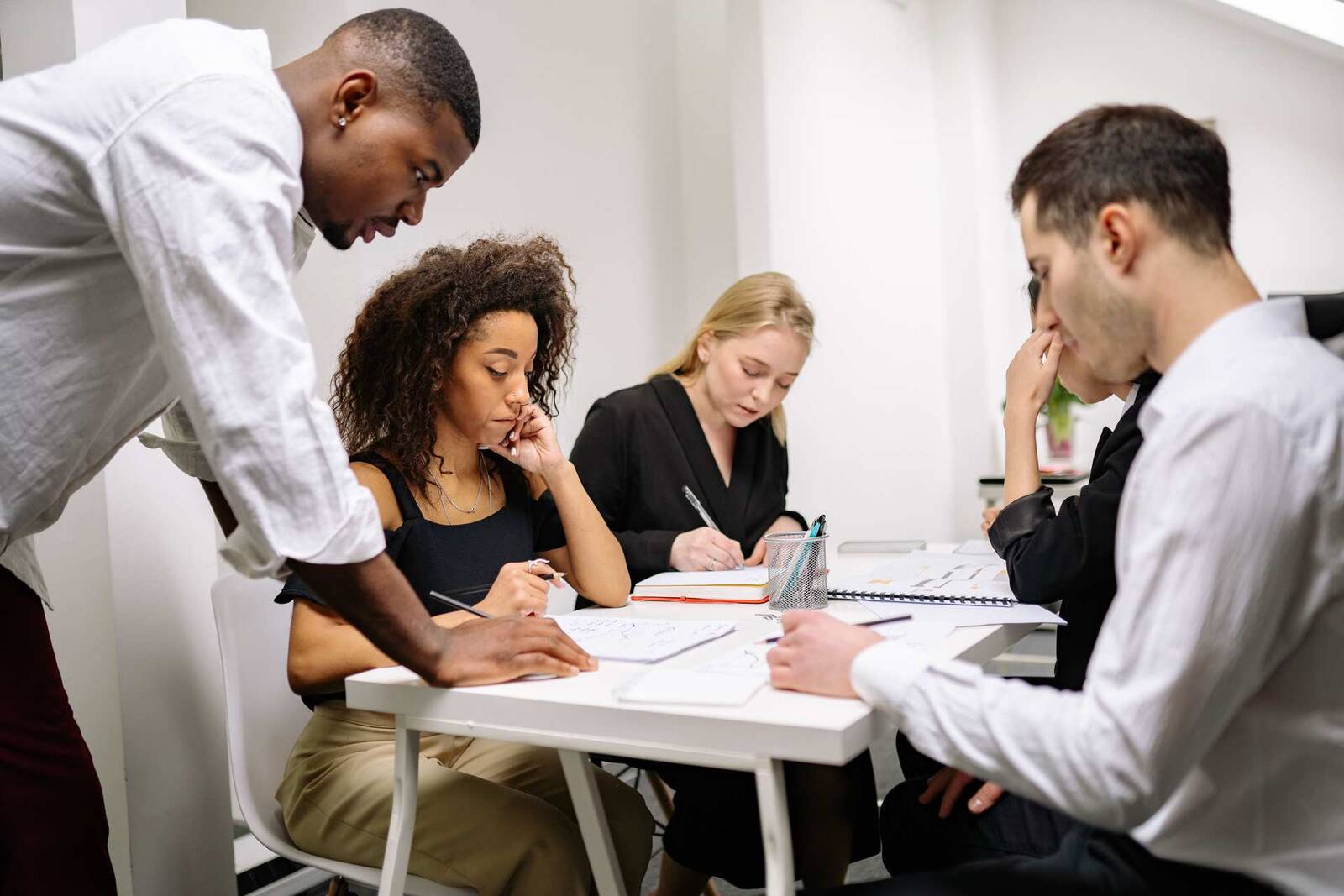 Five colleagues gather around a white table reviewing sketches and notes together, one standing and pointing