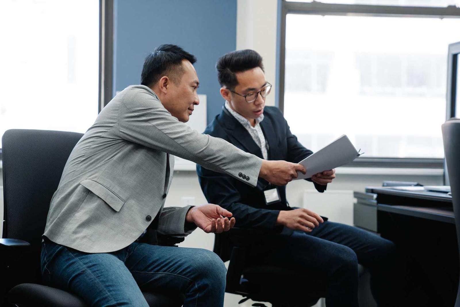 Two colleagues review a document together in an office, one gesturing while pointing at a page
