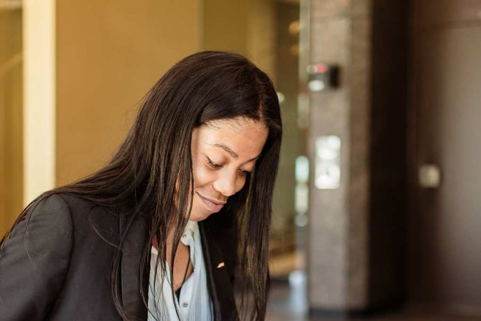 A woman in a blazer looks down in thought while seated in an office lobby