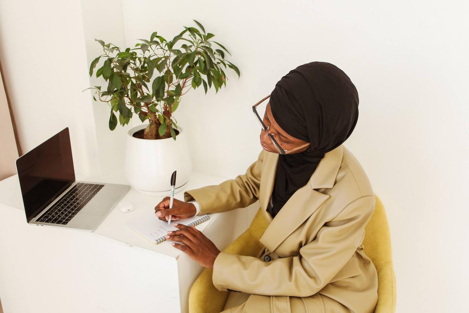 An overhead view of a woman in a headscarf and glasses writing in a notebook beside a laptop on a white desk, a plant nearby