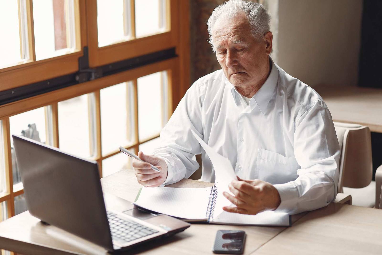 A senior man in a white shirt reviews handwritten notes beside an open laptop