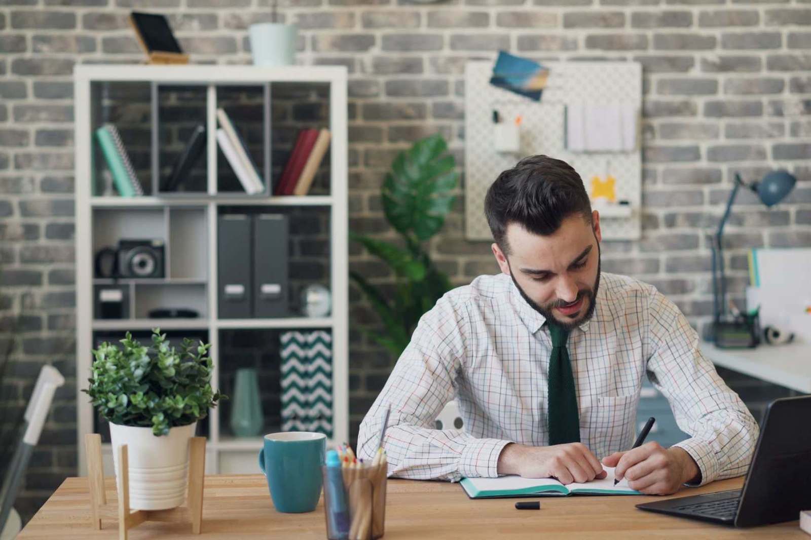 A man in a shirt and tie writes in a notebook at a desk beside an open laptop in a modern office