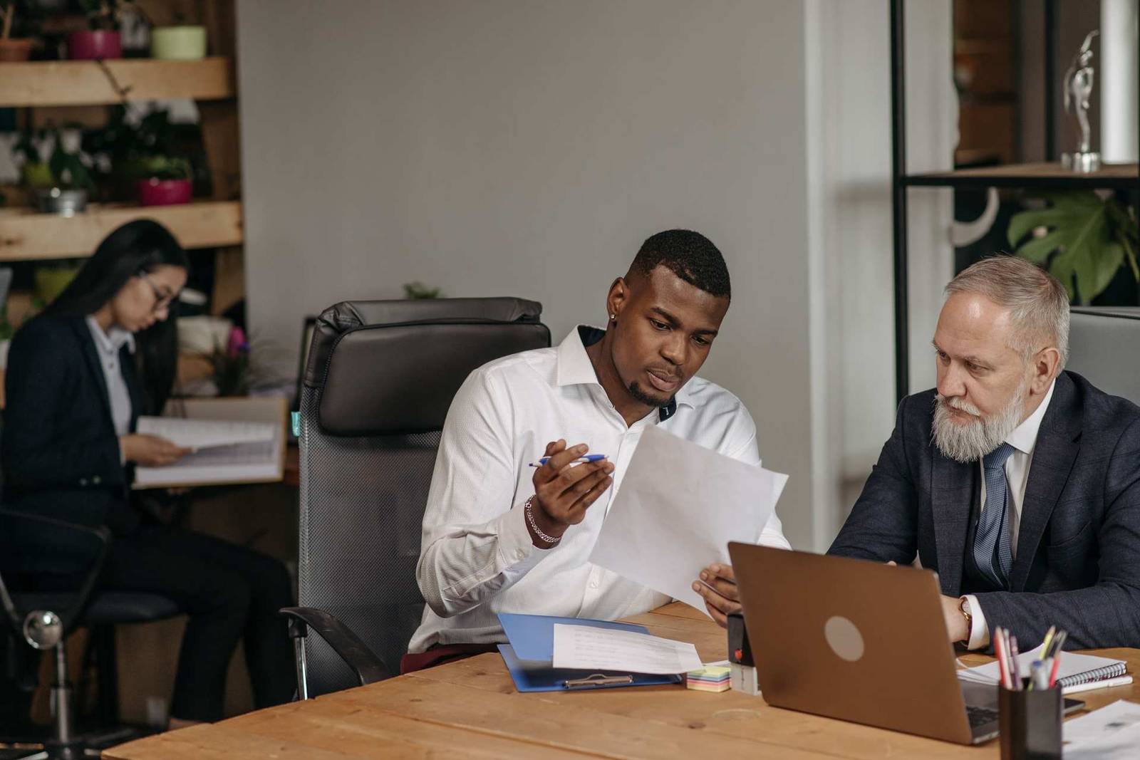 A senior colleague and a younger colleague review a printed document together at a desk, with a laptop open nearby