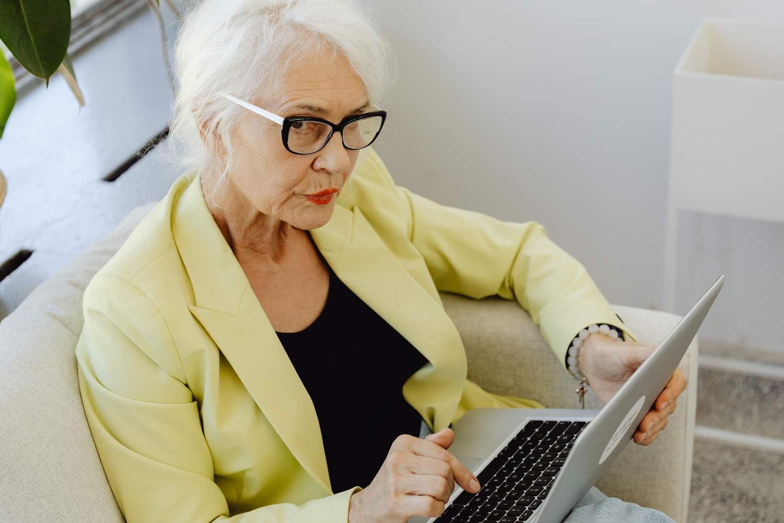 A woman in a yellow blazer and glasses looks closely at an open laptop while seated in an armchair