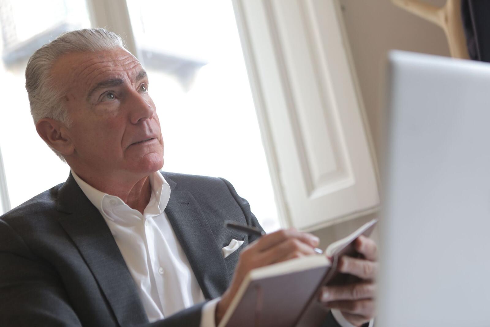 An older man in a grey blazer looks up in thought while writing in a notebook beside a laptop near a bright window