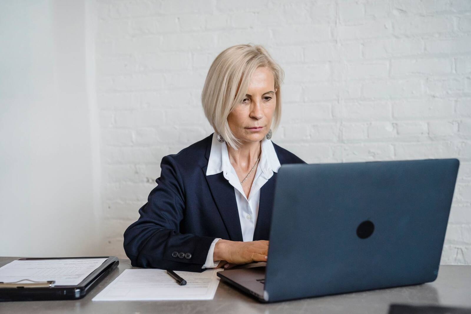 A woman in a navy blazer works intently at a laptop against a white brick wall