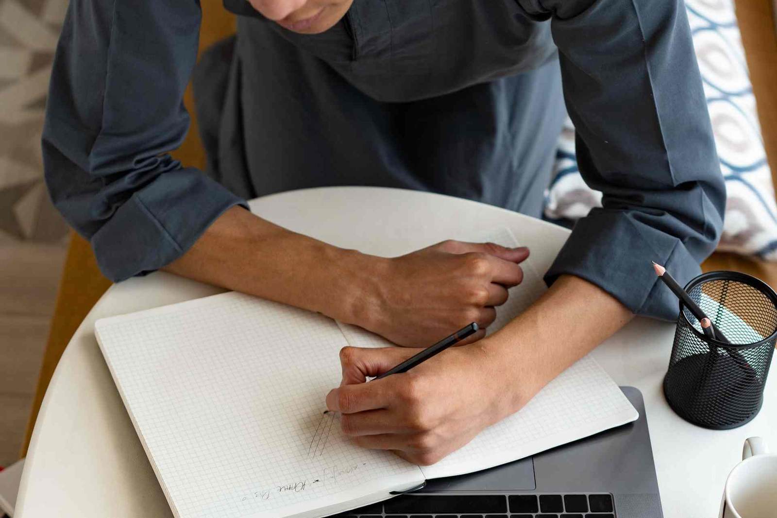 A close overhead view of a person's hands writing in a spiral notebook beside an open laptop and a cup of pencils