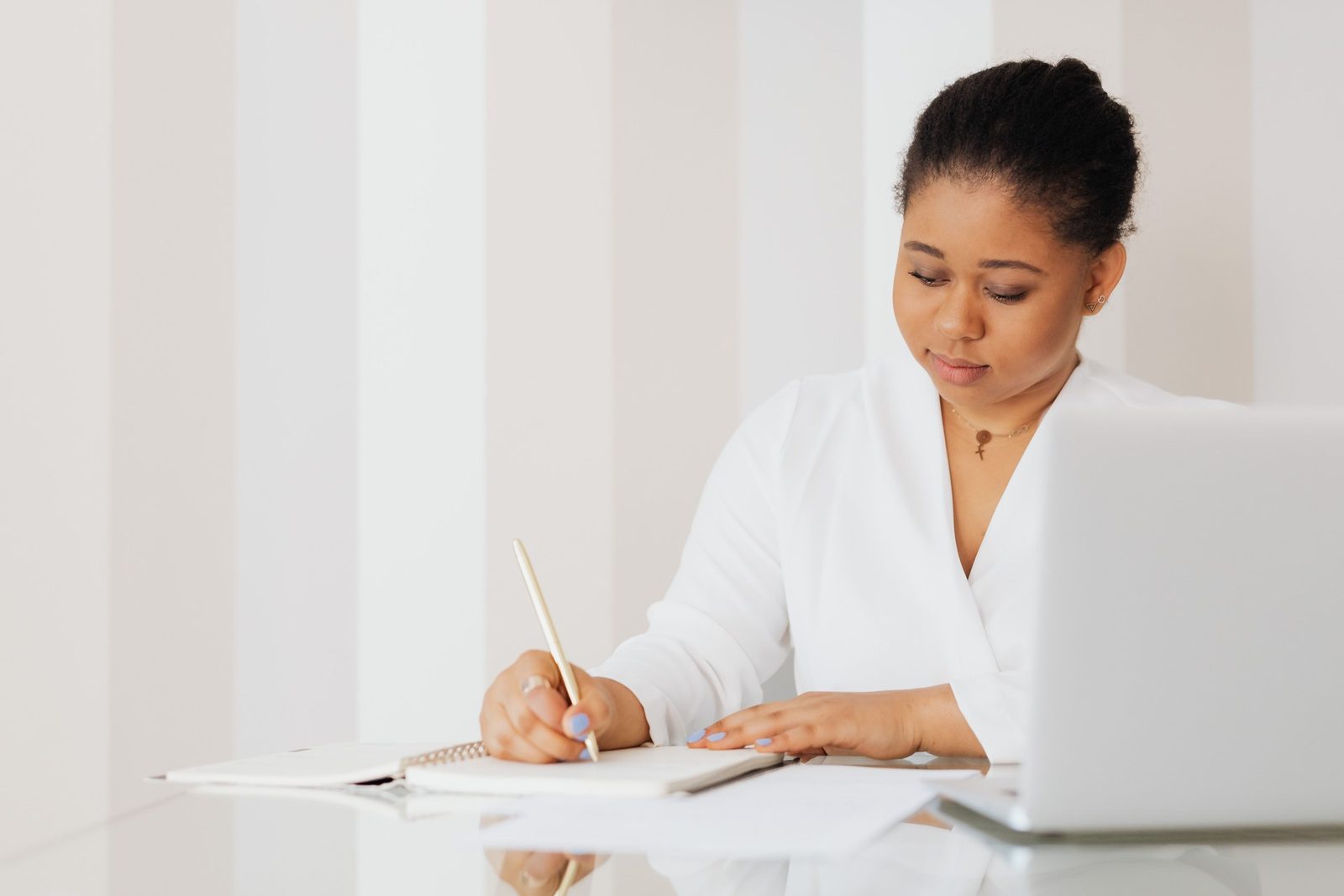 A woman writes closely in a notebook at a glass desk beside an open laptop