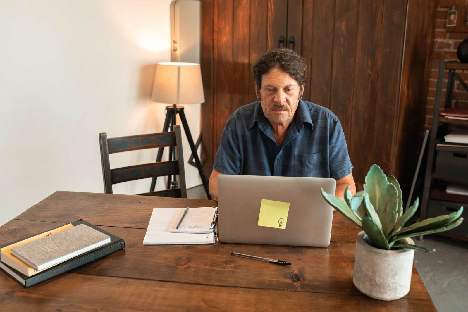 An older man reviews a laptop screen at a wooden desk with a notebook, pen, and plant beside him