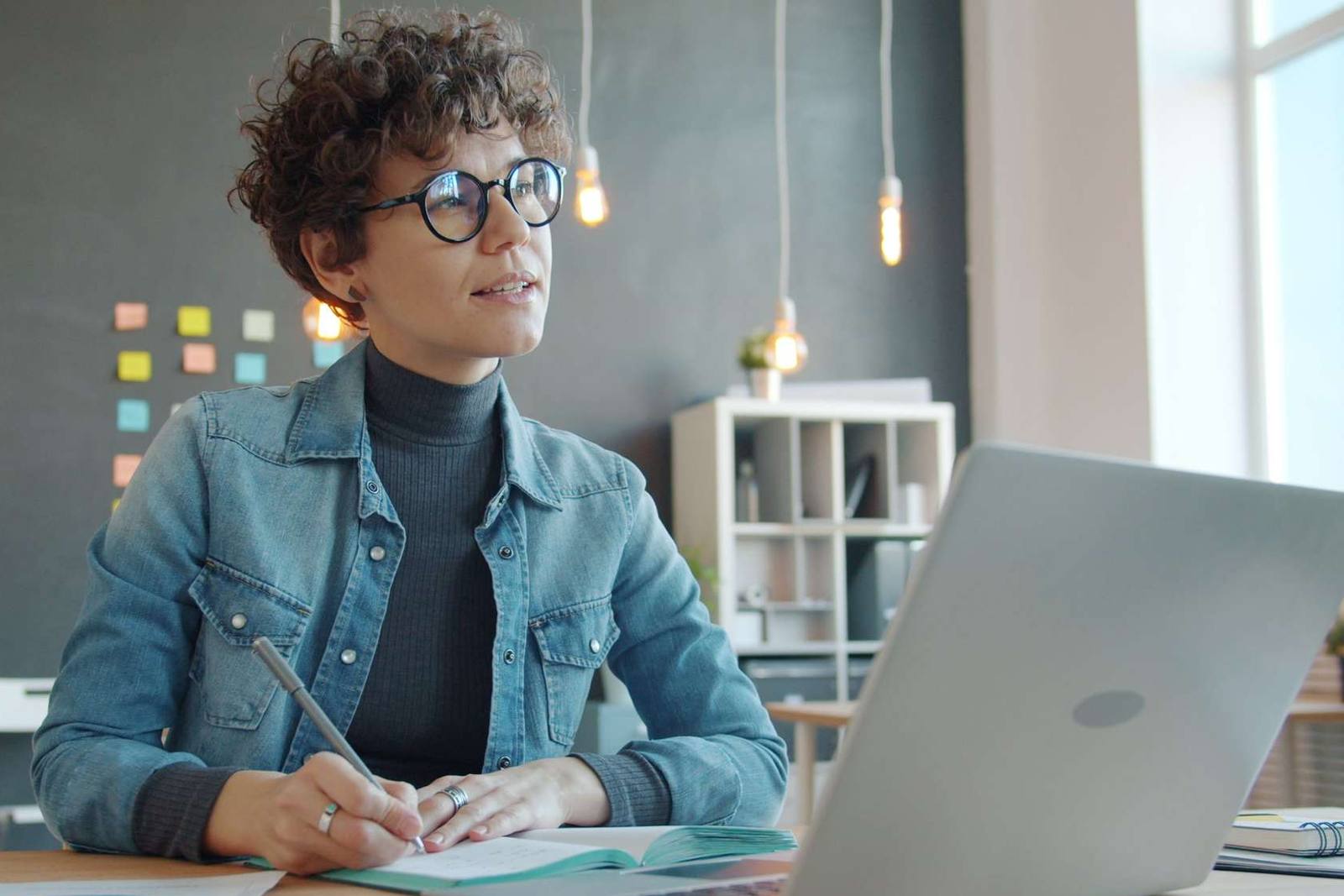 A woman in a denim jacket looks up in thought while writing in a notebook, laptop open in front of her