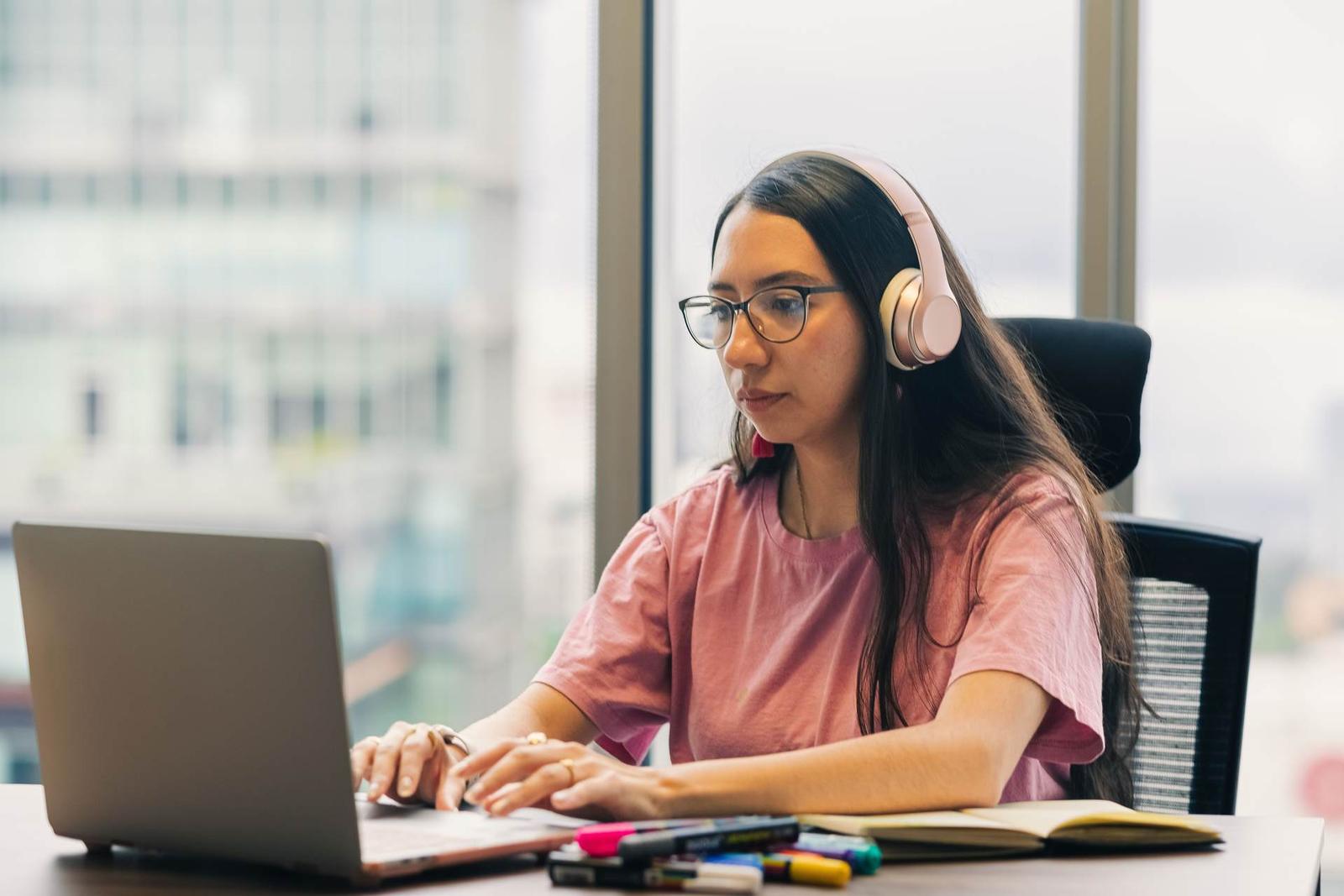 A woman wearing headphones reviews documents and types on a laptop at a desk with markers and an open notebook