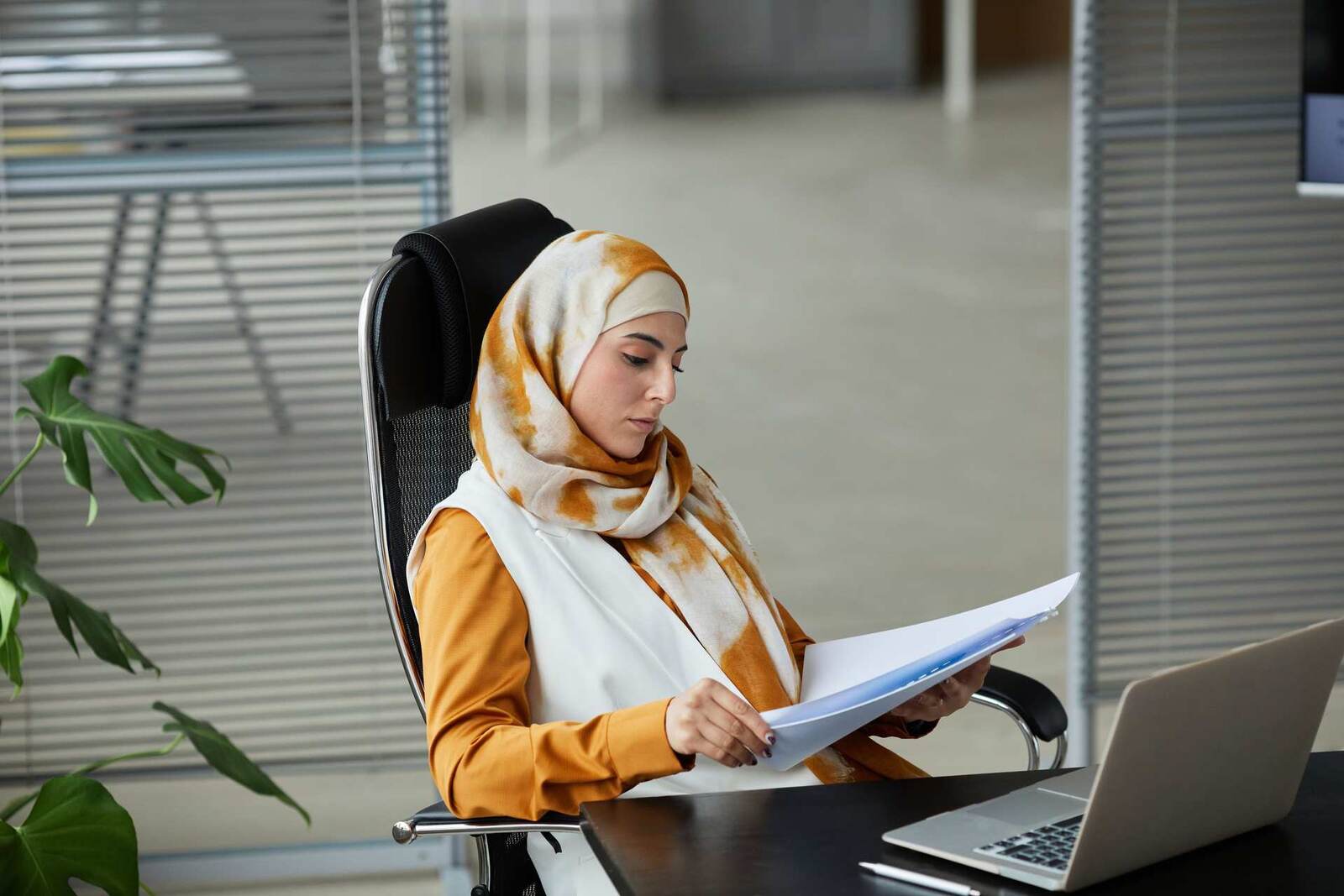 A woman in a patterned headscarf and white vest reads a sheaf of papers in an office chair, a laptop and plant nearby