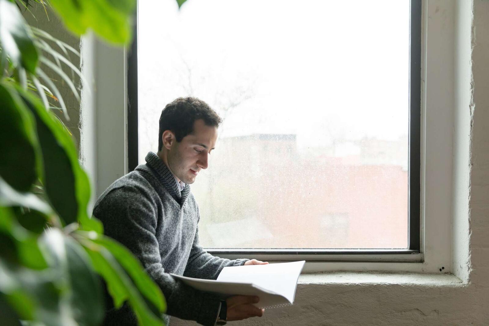 A man in a gray sweater reads a stack of papers by a tall window, with a houseplant in the foreground