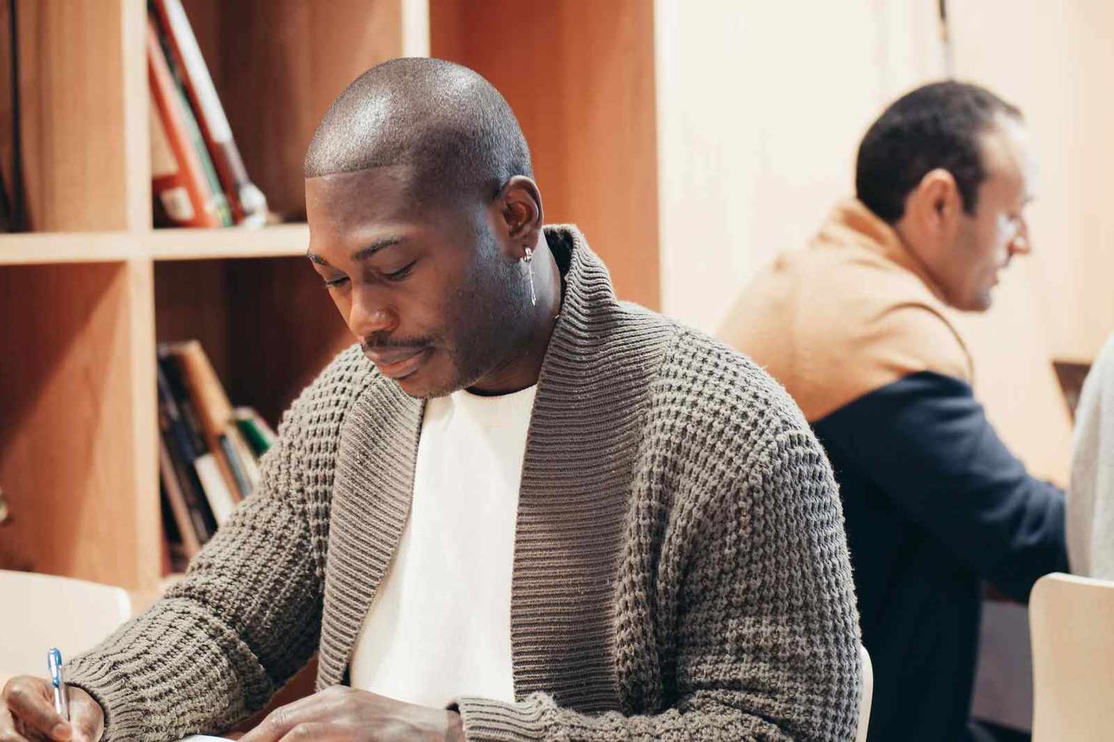 A man writes in a notebook in a book-lined room while colleagues work nearby