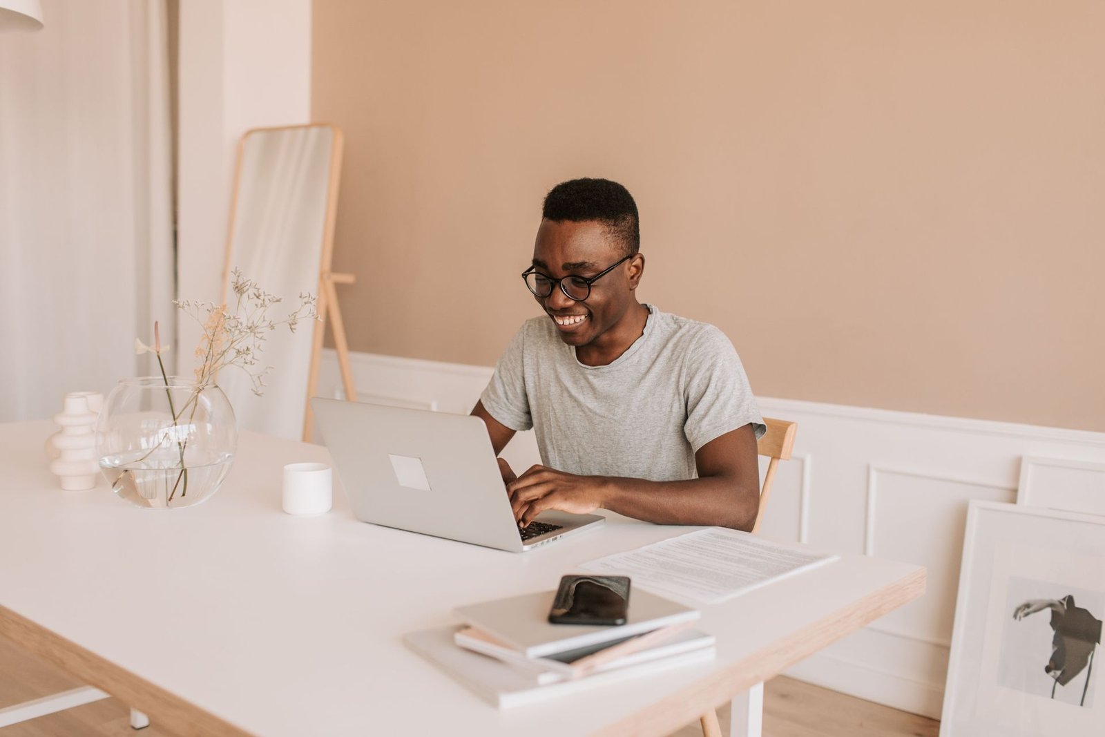 A man in glasses smiles while typing on a laptop at a bright desk in a warm-toned room