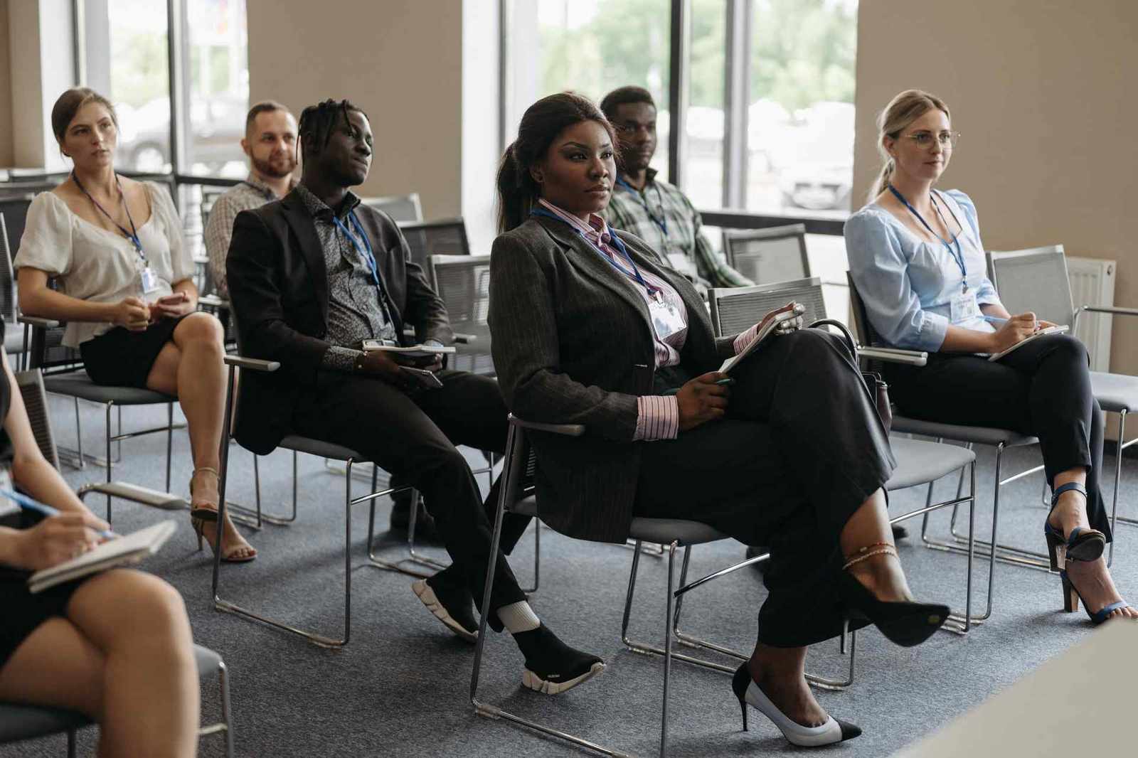 A row of seminar attendees wearing lanyards sit taking notes while listening in a bright meeting room