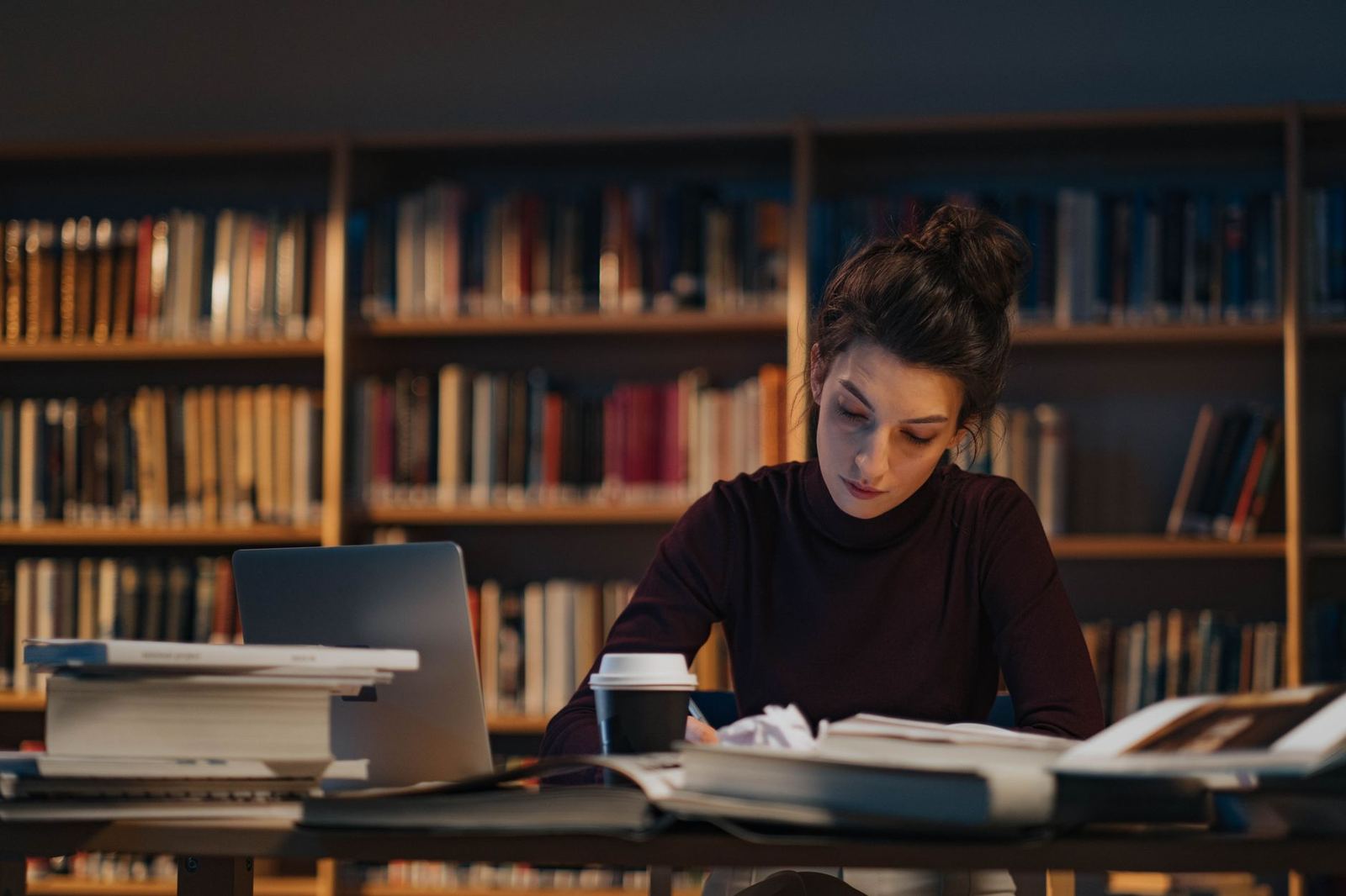 A woman studies late at night at a library table stacked with books and a laptop, rows of bookshelves behind her