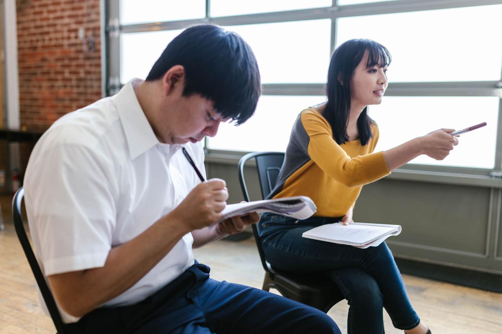 Two colleagues in a workshop setting, one writing notes on a pad and the other gesturing while speaking