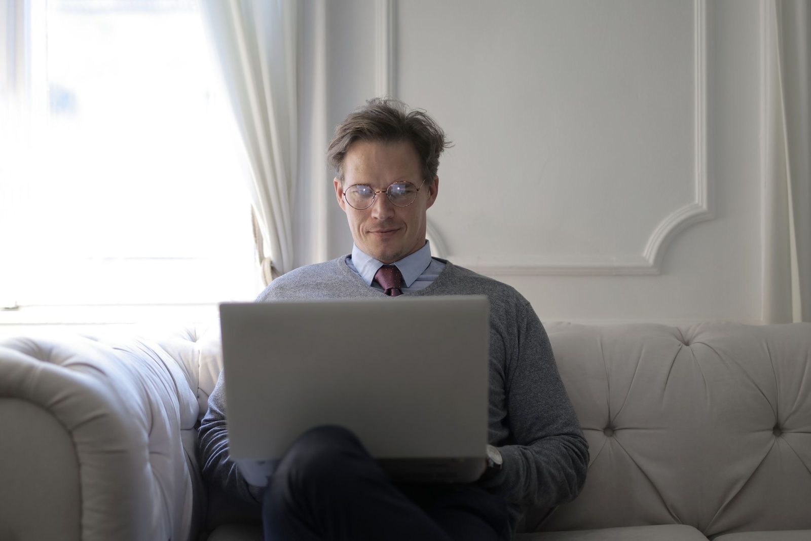 A man in glasses and a sweater and tie sits on a sofa working on a laptop, looking up in thought