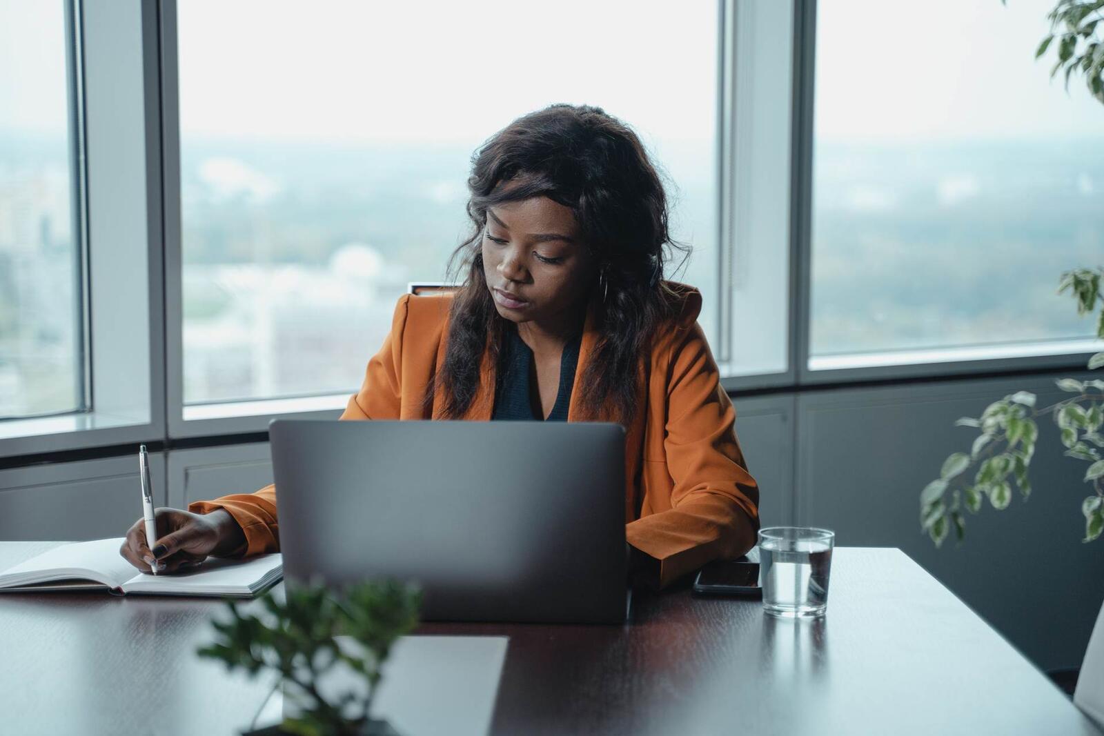 A woman in an orange blazer writes notes beside a laptop at a boardroom table, tall windows over the city behind her