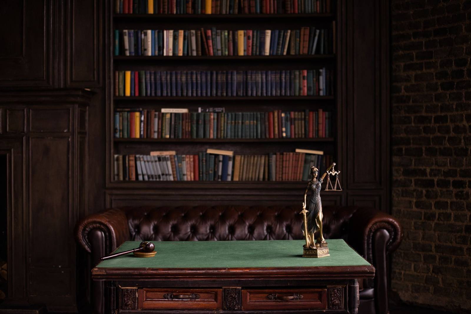 A gavel and a small blind-justice statuette holding scales, on a green desk pad in front of a dark wood bookshelf lined with law books